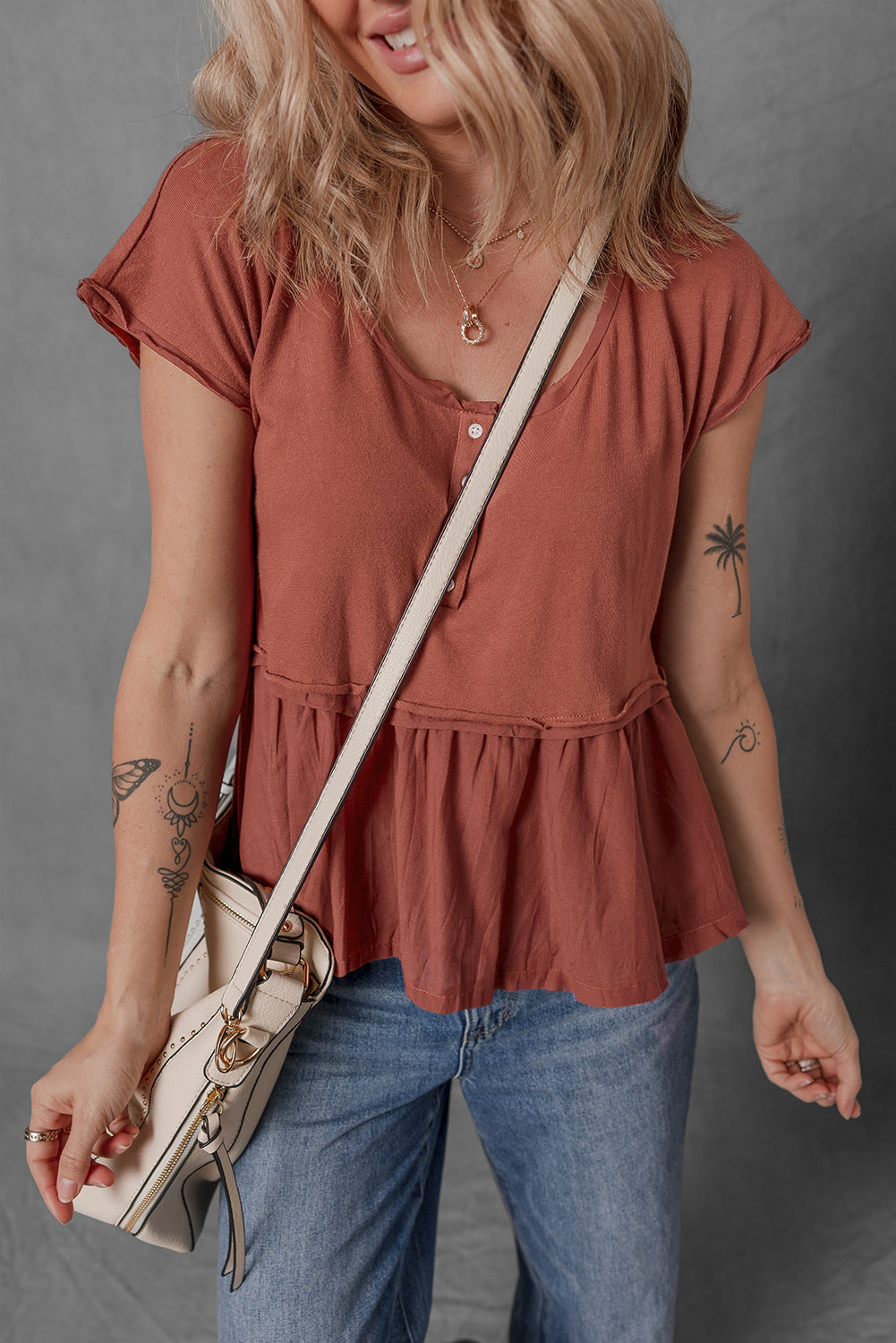 Woman wearing a rust-colored top and blue jeans, holding a beige handbag against a gray background